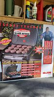 Photo of the George Foreman grill box showing front and side with branding, features, and marketing information including the image of George Foreman and cooked burger patties on the grill surface.