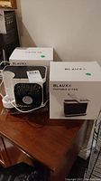 Three mini portable Blaux AC fans with one unboxed placed on wooden table next to two boxed units.