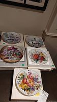 Photo showing five collector plates with floral designs arranged on boxes. Plates include round and square shapes, each with detailed colorful floral patterns. Certificates of Authenticity visible beneath some plates.