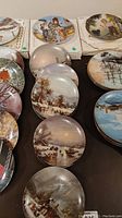 Photo showing 9 German collector plates arranged with visible winter theme scenes including frozen rivers, snow-covered cottages, and village people.