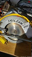 Close-up photo of Skilsaw circular saw showing metal blade guard, yellow power cord, and handwritten "Skilsaw" on the housing.