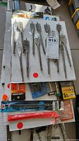 Photo of several flat wood drill bits (spade bits) mounted on white board, varying sizes, some loose, some in plastic packaging. Includes packaging with SpeedOut 2000 brand visible.