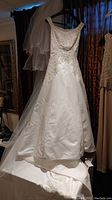 Full view of the white beaded wedding dress with veil hung behind it. Shows the front design and details of the fabric and beading.