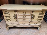 Front view of Asian-style chest showing light wood finish, metal corner braces, ornate handles, three central drawers, and two side doors.