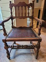 Front view of the wooden armchair showing carved backrest, armrests, and leather seat with nailhead trim.