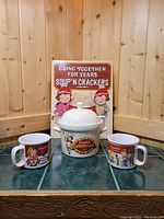 Photo showing a group of items: two ceramic Campbell's Kids soup mugs, a white ceramic crock pot with Campbell's Mock Turtle Soup label and kids artwork, and a vintage-style metal sign behind them.