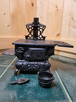 Front view of miniature Crescent cast iron stove with kettle and trivet on tile floor.