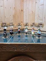 Full set of six Goebel wine glasses each with a unique hand-painted figurine stem, glass bowls etched with grapevine and gold trim, photographed on a glass top surface with wood background.