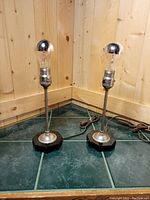 Two identical modern industrial style lamps with black bases and silver metal posts holding silver-tipped silver bowl light bulbs, placed on tiled floor against wooden panel wall.