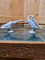 Two Lladro white porcelain swan figurines shown side by side on tiled floor against wooden panel background.