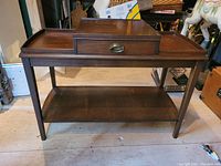Front view of Mersman wooden table showing the dovetailed drawer with brass handle and lower shelf, top surface with raised edges and central elevated panel.