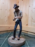 Front view of the painted cowboy sculpture standing on a round base in a wood-paneled corner, showing detailed face and hands counting coins.