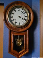 Full view of the wooden regulator school house clock hanging on a wall showing the wood grain, circular dial with Roman numerals, and brass pendulum behind glass door.