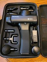 Photo of Sonic LifePro massage gun and seven interchangeable attachment heads placed in black foam insert inside carrying case, alongside power charger.