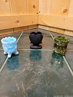 Photo showing all three vintage toothpick holders on a tiled floor corner: a blue glass holder with claw feet, a black holder with heart-shaped sides, and a green textured glass holder.
