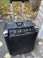 Black portable speaker with metal corner protectors, retractable handle, and front grille showing Alesis branding and control panel with microphone inputs and volume controls.