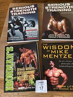 Photo showing four strength training books arranged on wooden floor with visible titles and cover images.