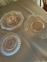 Three pink Depression glass plates arranged on a table showing differences in shape and size including a fluted round plate, an octagonal plate with handles, and a smaller scalloped round plate.