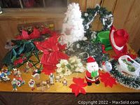 Top-down photo showing a small white artificial Christmas tree surrounded by red poinsettia bows, a Santa Claus figurine, greenery garlands with bows, and other assorted Christmas decorations and figurines on a wooden surface.