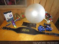 Overall lot view showing all exercise equipment items on a wooden table including stability ball, lifting belt, dumbbells, gloves and ab roller.