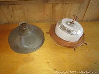 Two vintage glass light fixture shades displayed on a wooden surface against a wooden wall, showing one clear ribbed dome shade and one opaque decorative shade on a nautical ship wheel base.