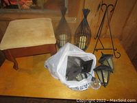 Photo showing two black metal teardrop candle holders with glass inserts, two black outdoor lantern lights, metal stand, clear glass orb, and a beige footstool on wooden table.