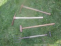 Photo showing two pick axes, one sledgehammer, and one garden fork laid on grass. Wooden handles and rusted metal heads visible.
