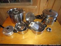 Photo showing front view of multiple large pots, pans, and stainless steel cookware including a grater and lids on wooden surface.