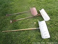 Three metal snow shovels laid on grass, showing variations in blade size and color and handle design.