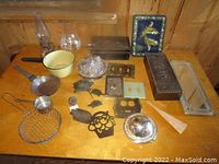 Various vintage kitchen and household items displayed on a wooden table including enamel saucepan, etched glass oil lamps, condiment set on silver tray, decorative tins, and metal utensils.