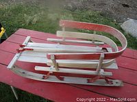 Side angle view of the wooden children's sleigh showing the red metal runners, curved wooden handle, and natural wood slatted base.