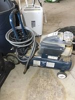 Side view showing blue air compressor tank with attached handle and wheels, coiled hose wrapped around handle, visible wear.