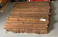Wood balusters stacked on the floor, rectangular in shape with a medium brown stain, showing some knots and natural wood grain.