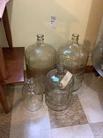Overview of three large glass carboys with ribbed horizontal bands and one smaller smooth glass jug grouped together on floor.