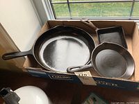 Three cast iron pans in a cardboard box by a window. One large round skillet and one square skillet and one round griddle pan visible.