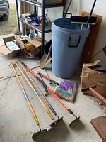 Wide view of the large Rubbermaid blue garbage pail, four garden hoes laid on the floor, broom, shovel, and caddy with tools around.