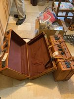 Two worn brown leather-like hard cases, one open showing brown fabric interior with pocket, the other closed, placed on floor with visible wear on exterior.