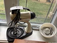 Vintage Sunbeam Mixmaster electric stand mixer with beaters and juicer attachment on side, placed on a black triangular base, near a window.