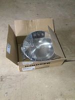 Open cardboard box containing a stainless steel vanity sink partially wrapped in protective plastic.