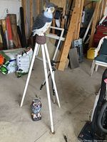 Full view of wooden tripod standing open with large plastic owl screwed on top and smaller owl figurine at base in a garage workspace.
