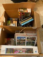 Two boxes filled with multiple jigsaw puzzle sets mostly complete in their original boxes and the boxed set of The Hunger Games Trilogy books.