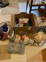 Box containing sewing accessories shown with multiple items around it on floor.