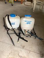 Pair of white backpack sprayers on concrete floor, showing BE and SP Systems logos with attached straps and wands.