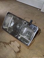 Photo showing the stainless steel kitchen sink with one large and one smaller basin, attached piping visible, placed on a cardboard box on a concrete floor.