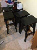 Set of six black wooden bar stools viewed from above and side, showing rectangular seats and square legs with horizontal footrests.