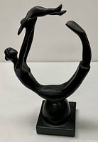 Side-angle view of black artistic statue of mother lying back lifting child figure above her, showing statue curved to form a circular arch, on square base.