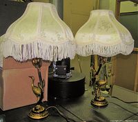Pair of gilt metal Art Nouveau style table lamps with fabric fringed shades placed on a table showing full view of lamps.