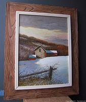 Front view of framed oil painting depicting barn in a winter landscape with snow field, fence and hillside background.