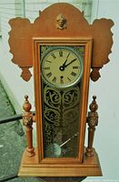 Front view of wooden gingerbread clock showing lion's head and decorative glass door with pendulum visible.