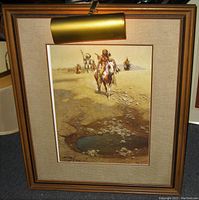 Framed art print with picture light switched on showing Indigenous braves on horseback near a desert water hole, signed McCarthy 1985.